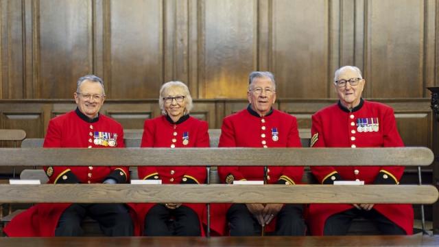 Four Chelsea Pensioners wearing scarlet uniforms sit together on a wooden bench inside the Wren Chapel. They face the camera with service medals displayed on their coats, set against wood panelled walls and historic seating.