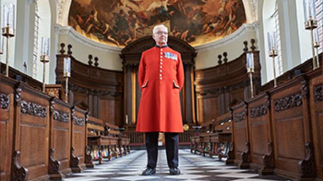 A Chelsea Pensioner, dressed in his iconic scarlet uniform, stands within a grand chapel with dark wooden pews on either side of a chequered black and white aisle. Leading to a wooden altar with a painted fresco above