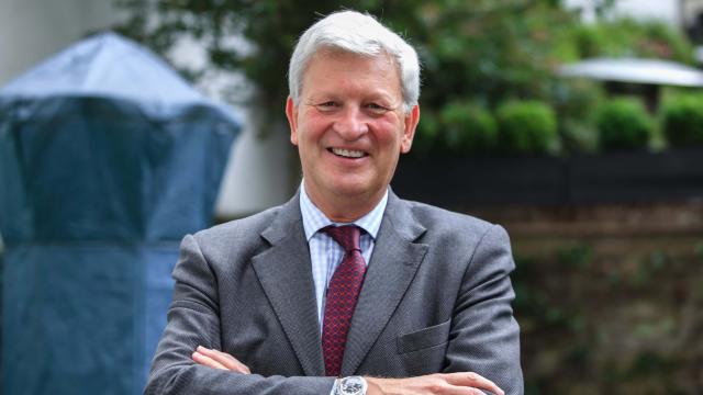 Sir Simon Mayall, a man stands with his arms crossed and a large welcoming smile on his face outdoors. His arms are folded in front of him and wears a grey suit with burgundy tie.