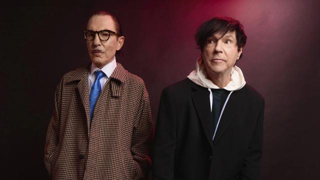 Two men stand side by side against a dark studio backdrop with subtle red lighting. One wears a patterned overcoat, glasses, and a tie, looking down thoughtfully, while the other wears a black suit layered over a light hooded top, looking off to the side. The image has a stylised, dramatic promotional feel.