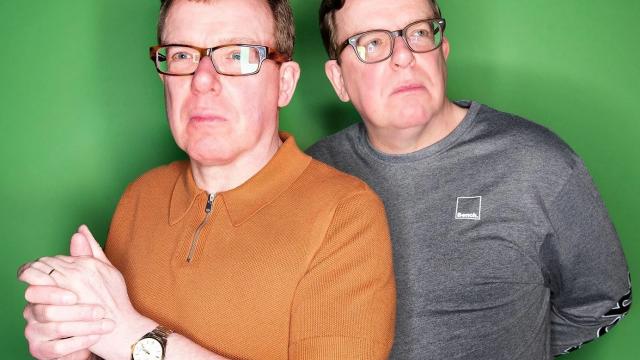 Two middle-aged men stand against a bright green background, both wearing glasses. One wears a short-sleeved rust-coloured polo shirt and clasps his hands, while the other stands slightly behind in a grey long-sleeved top. Both look off to the side with thoughtful, slightly serious expressions.