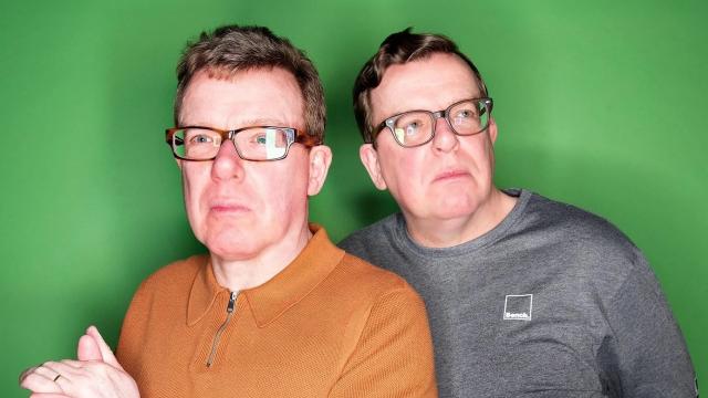 Two middle-aged men stand against a bright green background, both wearing glasses. One wears a short-sleeved rust-coloured polo shirt and clasps his hands, while the other stands slightly behind in a grey long-sleeved top. Both look off to the side with thoughtful, slightly serious expressions.