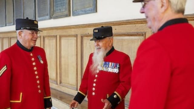 Three Chelsea Pensioners in traditional scarlet uniforms stand together indoors, talking and smiling. They wear black RH caps and service medals. The setting features wooden wall panelling and a light, historic interior at the Royal Hospital Chelsea.