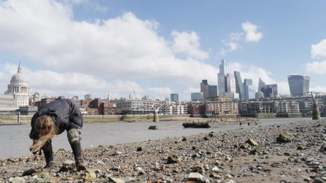 A person bends down on a rocky riverbank, examining the ground near the water’s edge. Across the river, the London skyline is visible, including St Paul’s Cathedral and modern skyscrapers. The scene is set under a bright sky with scattered clouds, contrasting natural textures with the urban cityscape.