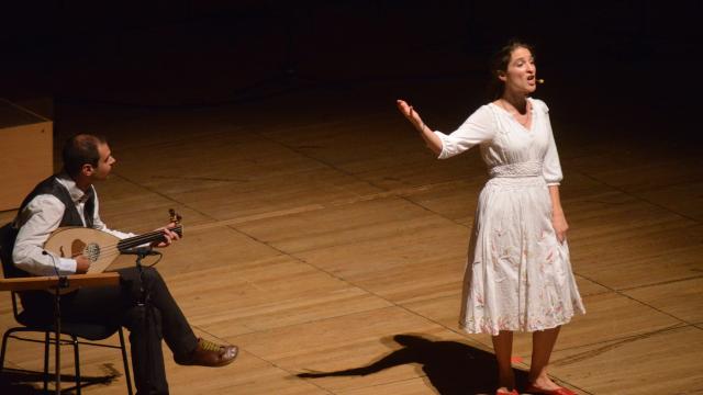 A lady stands in white dress stands on a wooden stage - one arm raised as she sings. She is basked in warm light as a well dressed man sits to the left of the stage playing a loot.