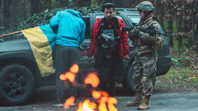 A tense scene on a forest road where a civilian in a red jacket stands beside a military vehicle partially covered with camouflage netting. A Ukrainian flag is draped over the vehicle. An armed soldier in camouflage stands nearby, while a small fire burns in the foreground, casting warm light against the dark woodland setting.
