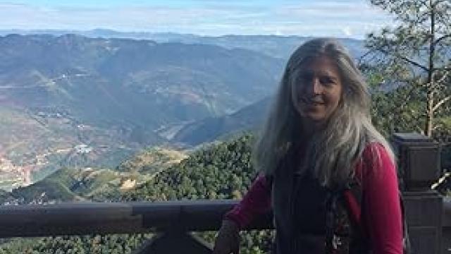 A woman stands at a wooden viewpoint overlooking a wide mountain valley. She wears a red long-sleeved top and a dark vest, smiling towards the camera. Rolling hills, forests, and distant mountains stretch across the background under a bright sky.