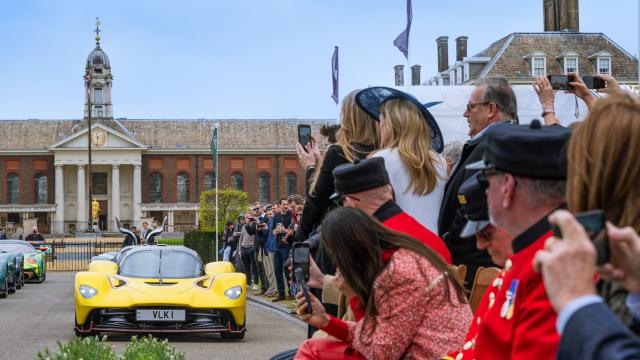 A yellow sports car stands on the south grounds of the Royal Hospital Chelsea, with the iconic building in the background and numerous spectators, including the Chelsea Pensioners dressed in their iconic scarlet uniforms, arranged alongside the right of the car.