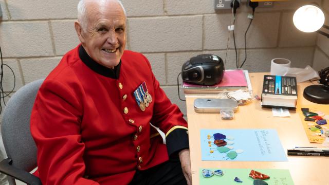 A Chelsea Pensioner wearing a scarlet uniform with service medals sits at a desk smiling towards the camera. In front of him are colourful pieces of glass and artwork arranged on paper, alongside craft tools and a desk lamp. The setting appears to be a workshop or studio space, suggesting a relaxed and creative activity.