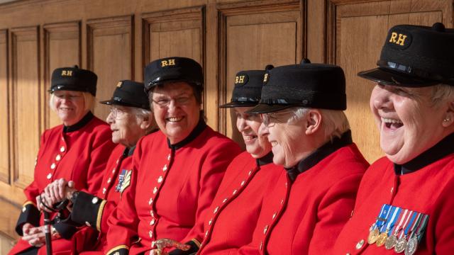A group of Chelsea Pensioners sit side by side on a wooden bench indoors, wearing their scarlet uniforms and RH hats. They are smiling and laughing together, with medals visible on their coats. Several hold walking sticks, and the scene conveys companionship, pride, and shared humour.