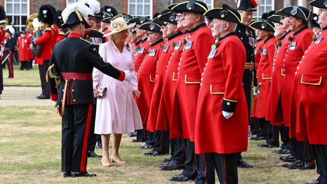Queen Camilla walks along a line of Chelsea Pensioners in scarlet uniforms during a ceremonial parade at the Royal Hospital Chelsea, accompanied by military officers in full dress uniform as she speaks with participants.