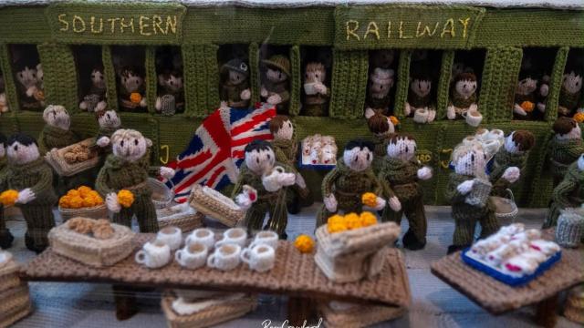 Crocheted diorama of soldiers boarding a Southern Railway carriage, greeted with tea, buns and marigolds, with a Union Jack flag, part of The Longest Yarn WWII display.