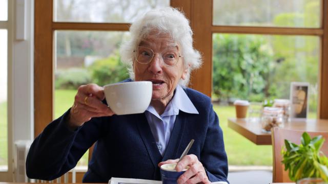 An Image of Chelsea Pensioner Marjorie holding up a cup of tea at the cafe at Royal Hospital Chelsea